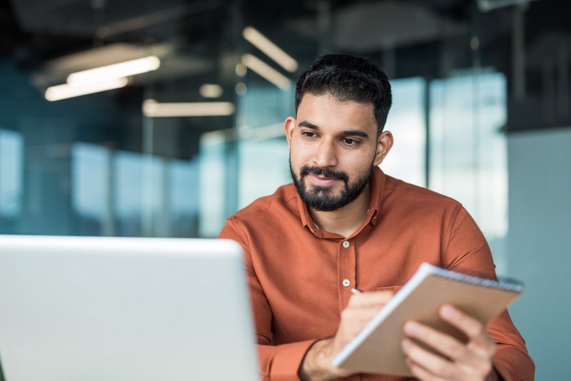 Person writing on notepad while looking at computer screen.