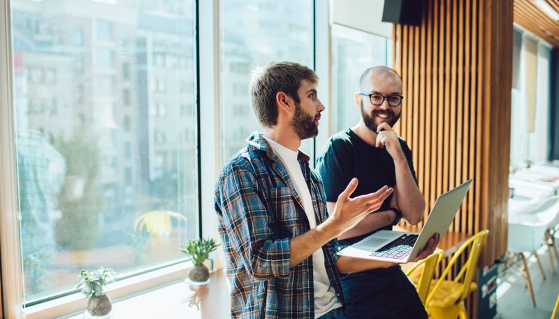 Two people talking in a modern office while holding a laptop.