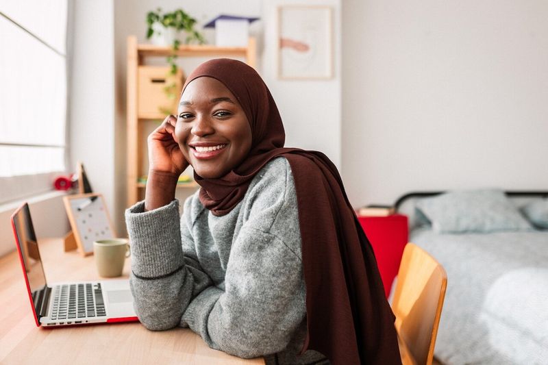 Student sitting with laptop at study desk in bedroom.